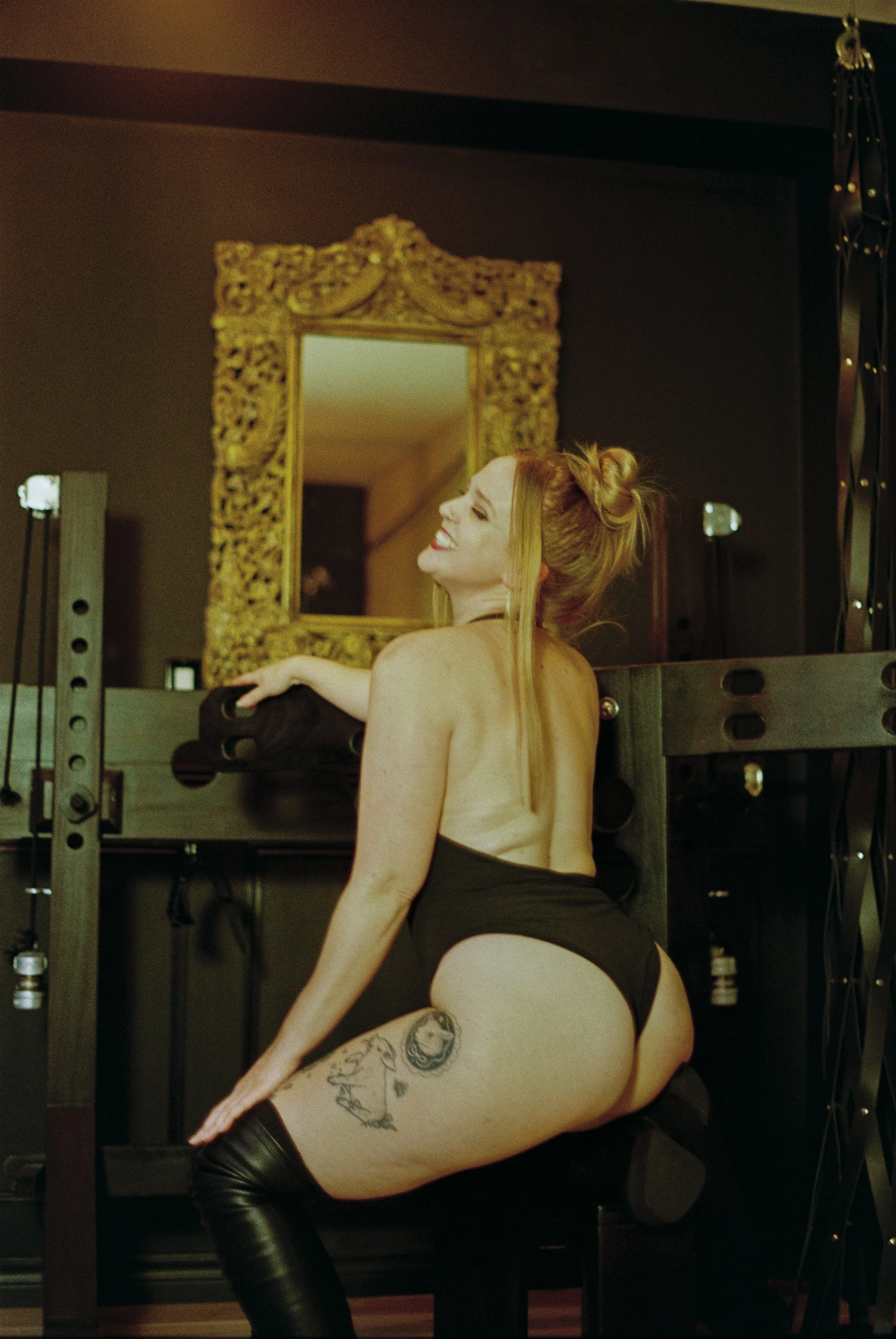A woman with long red hair styled in a messy bun, smiling with eyes closed, wearing black lingerie and thigh-high boots, sitting on a bench in a room with dark walls, a gold ornate mirror, and fitness equipment nearby.