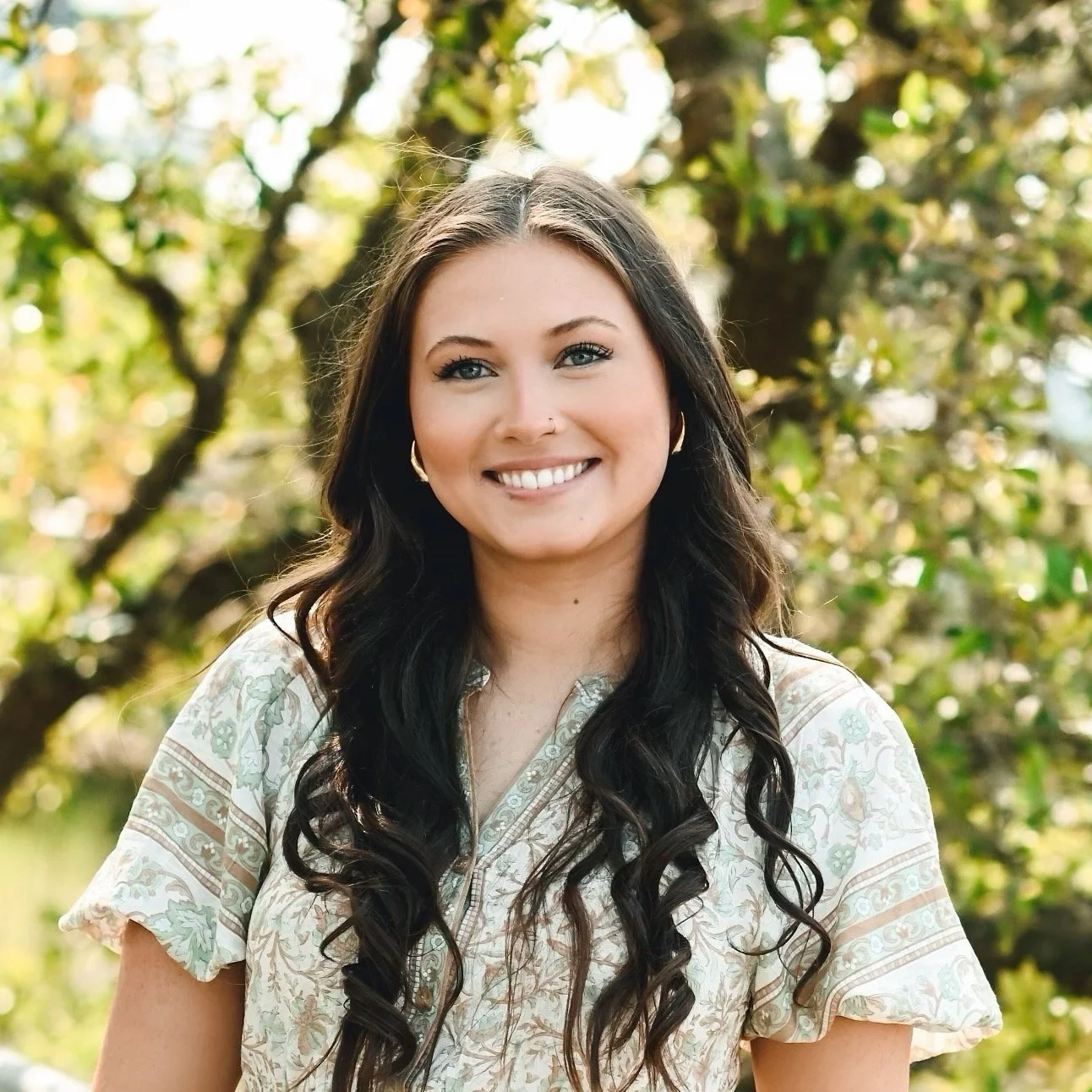 A woman with long dark wavy hair and a nose piercing, smiling outdoors with trees in the background.