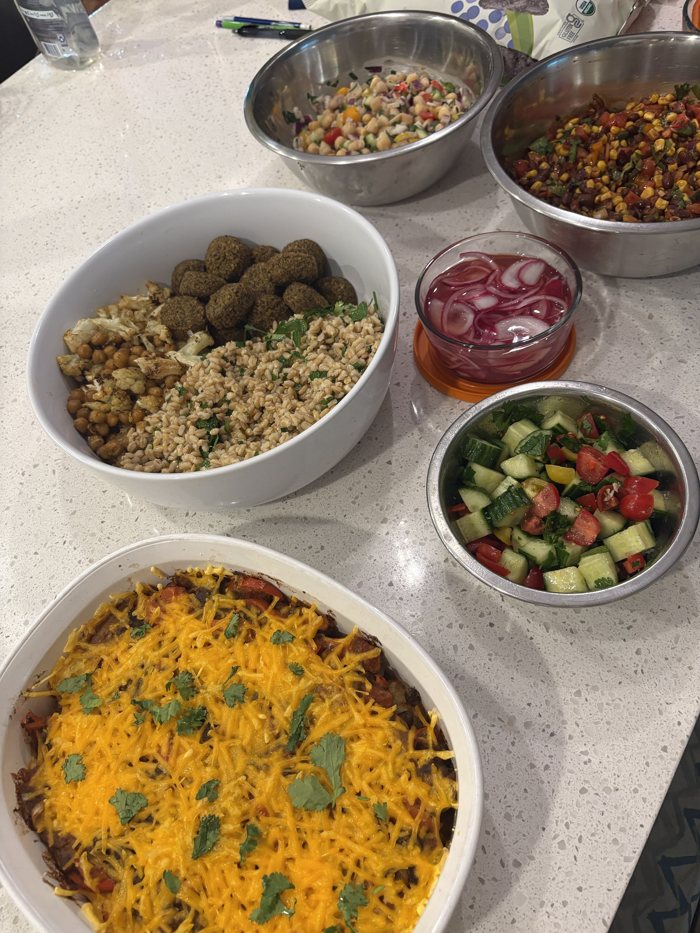 A spread of various dishes on a white countertop, including a baked casserole topped with shredded cheese and cilantro, a bowl of falafel balls with chopped chickpeas, diced vegetables salad, a bowl of mixed bean salad, a cucumber and tomato salad, and a glass of pink drink with ice.