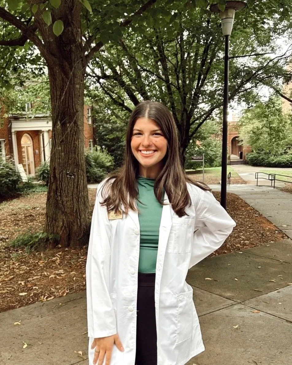 A woman with dark brown hair who's smiling, and wearing a green shirt and a white coat, outdoors with trees and brick buildings in the background.