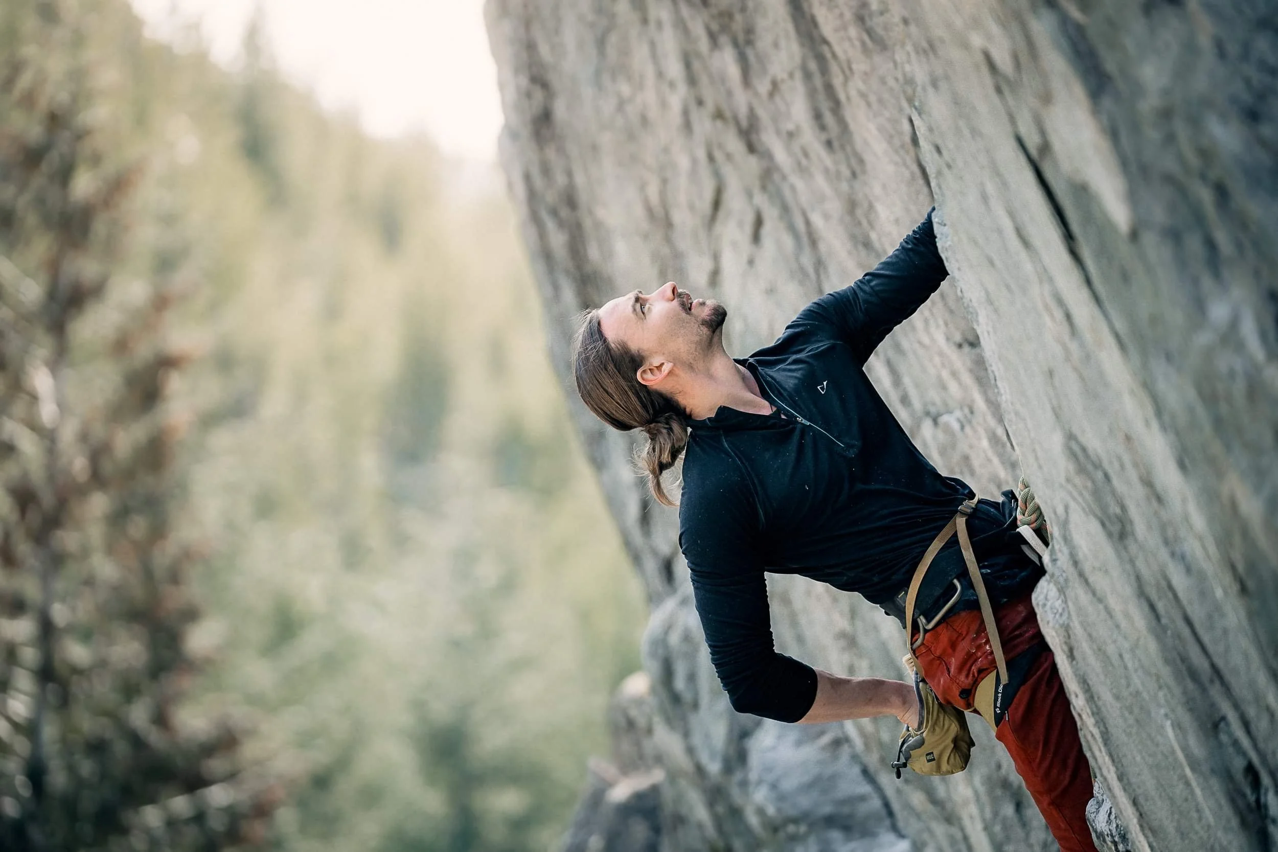 A man rock climbing on a vertical cliff face, wearing outdoor gear, with a background of blurred trees and sky.
