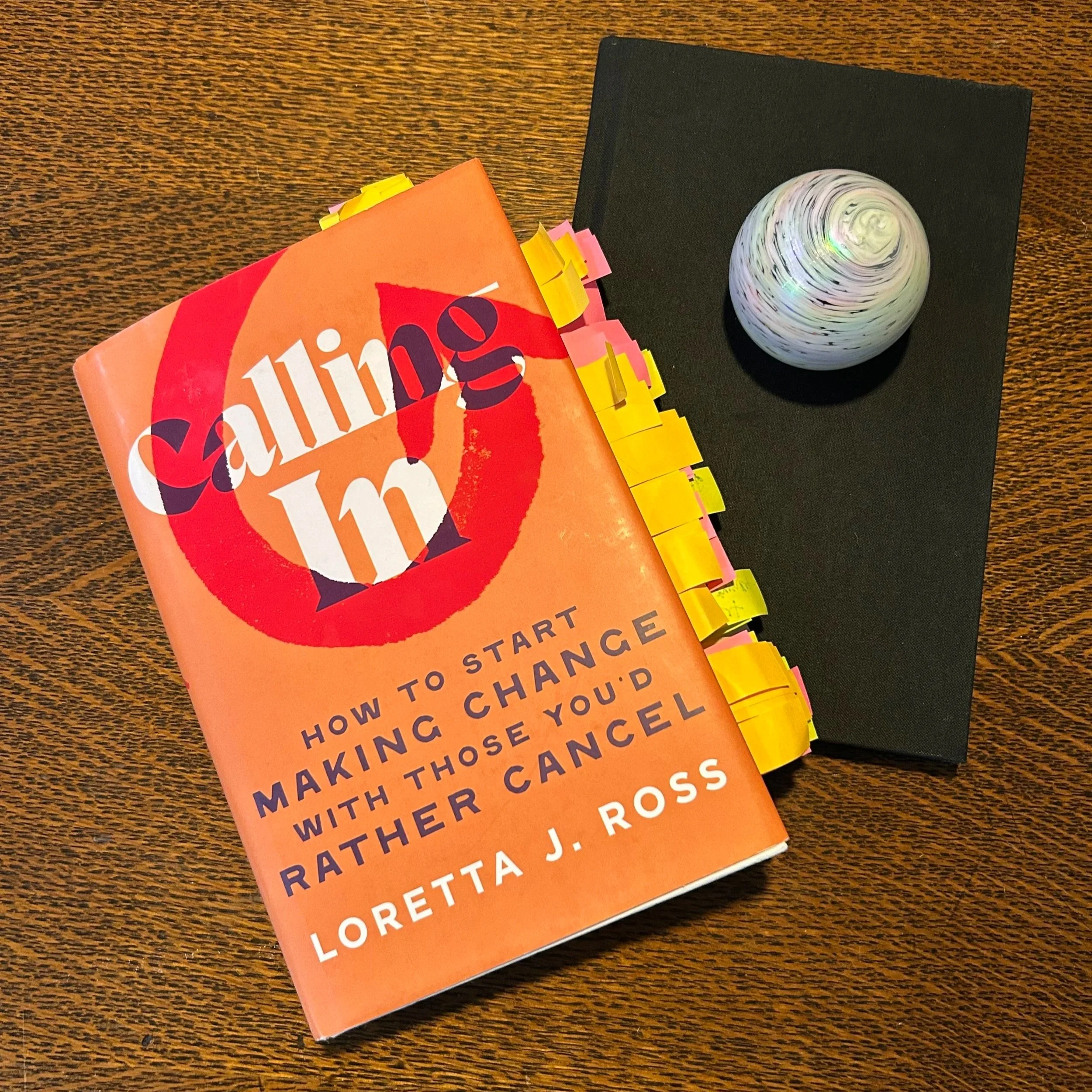 Photo of hardcover book, "Calling In" by Loretta J. Ross. Book is tabbed with dozens of colorful flags. Next to book is black journal with white swirly paperweight on top.