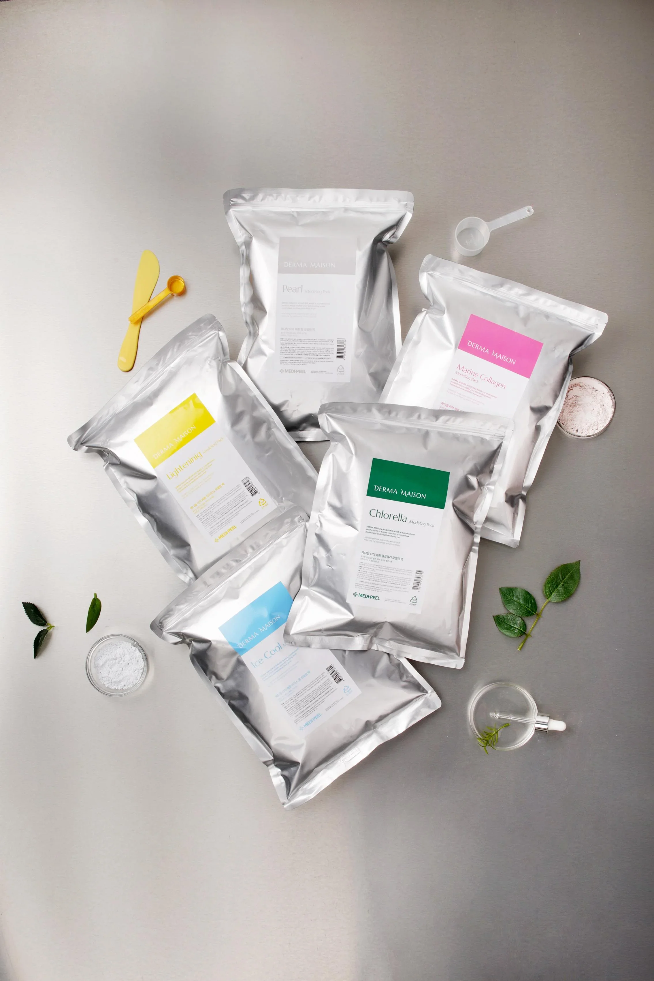 Multiple silver pouches of Derma Maison skincare products on a grey surface, with small bowls of powder, a measuring scoop, a yellow spatula, green leaves, and a small glass container with a dropper.
