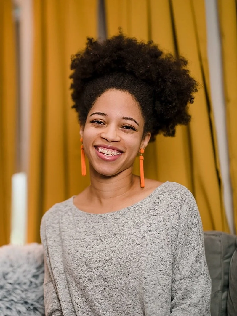 A Black woman with brown natural hair wearing a light grey top with dangling orange earrings, sitting on a grey couch next to a fuzzy grey pillow in front of yellow curtains.