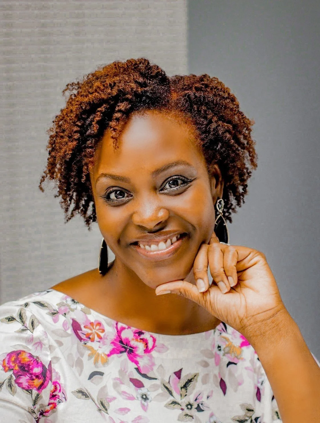 A Black woman with brown natural hair wearing a floral dress, sitting in front of a grey wall.