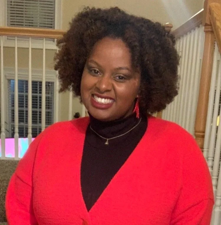 A Black woman with brown natural hair wearing a black turtleneck under a red cardigan, standing in front of a wooden railing.