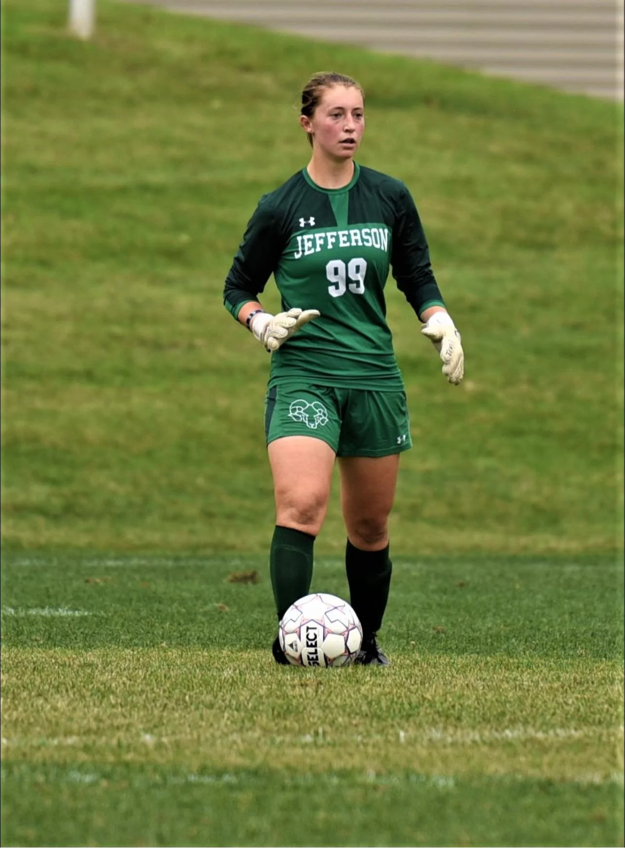 Soccer player in green Jefferson uniform with the number 99, wearing gloves, standing on grass field with soccer ball.