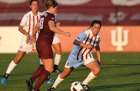 Soccer players in action on a field, wearing uniforms with stripes and solid colors. One player is controlling the ball, while others look on. Sunlight creates strong shadows on the grass.