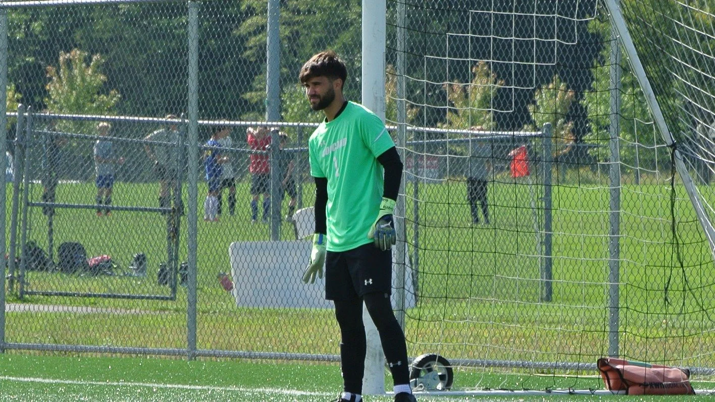 Soccer goalkeeper standing in front of the goal during a game with a chain link fence and other people in the background.