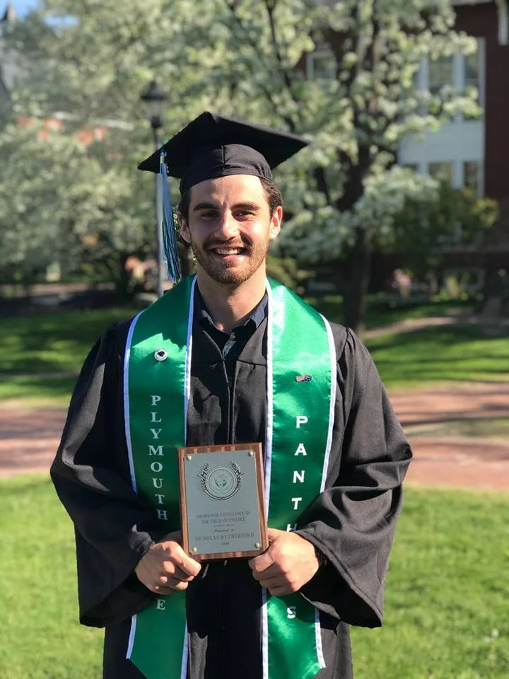 A graduate wearing a cap and gown stands outdoors holding a plaque. The gown is black with green and white sashes that say 'Plymouth Panthers.' The background features blooming trees and a grassy area.