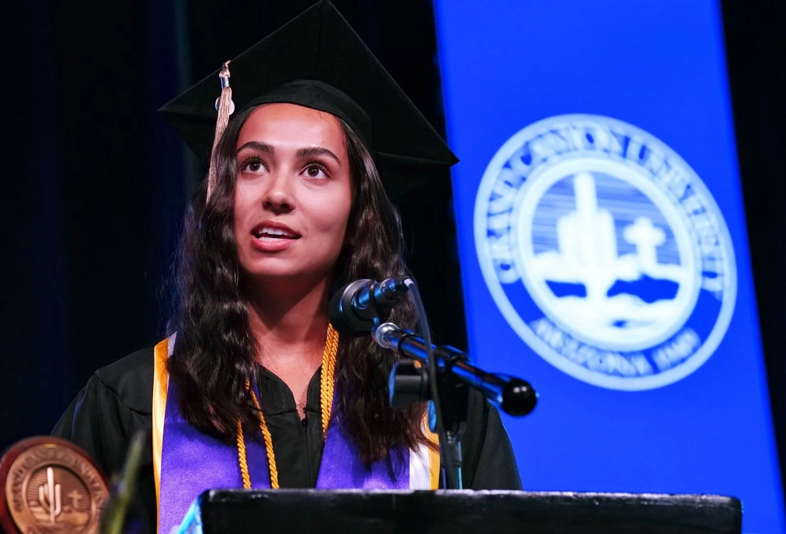 A person in a graduation cap and gown speaking at a podium, with a university emblem on a blue background behind them.