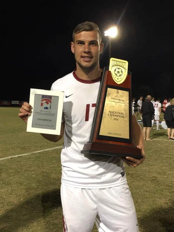 Soccer player holding NAIA National Champion trophy and certificate on a soccer field.