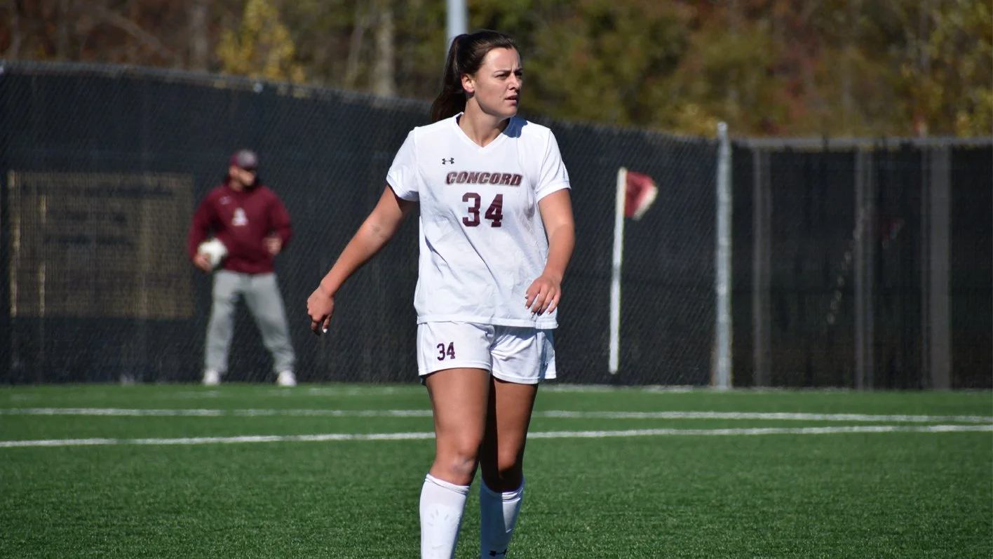 Female soccer player on field wearing a white uniform, jersey number 34, with "Concord" on the shirt, and a person in the background with a ball.