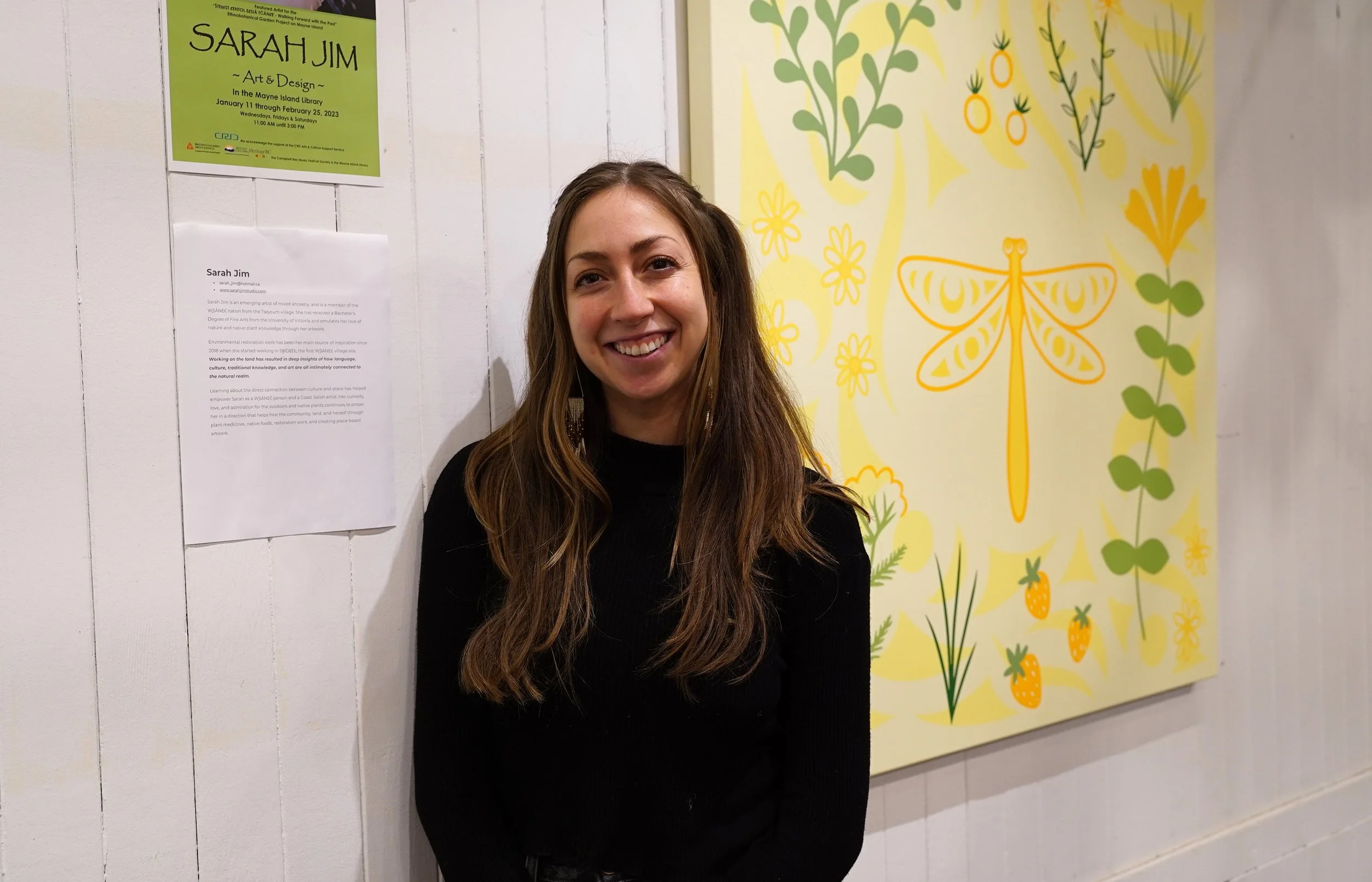 Portrait of Sarah Jim, a person with long brown hair, smiling in front of her artwork