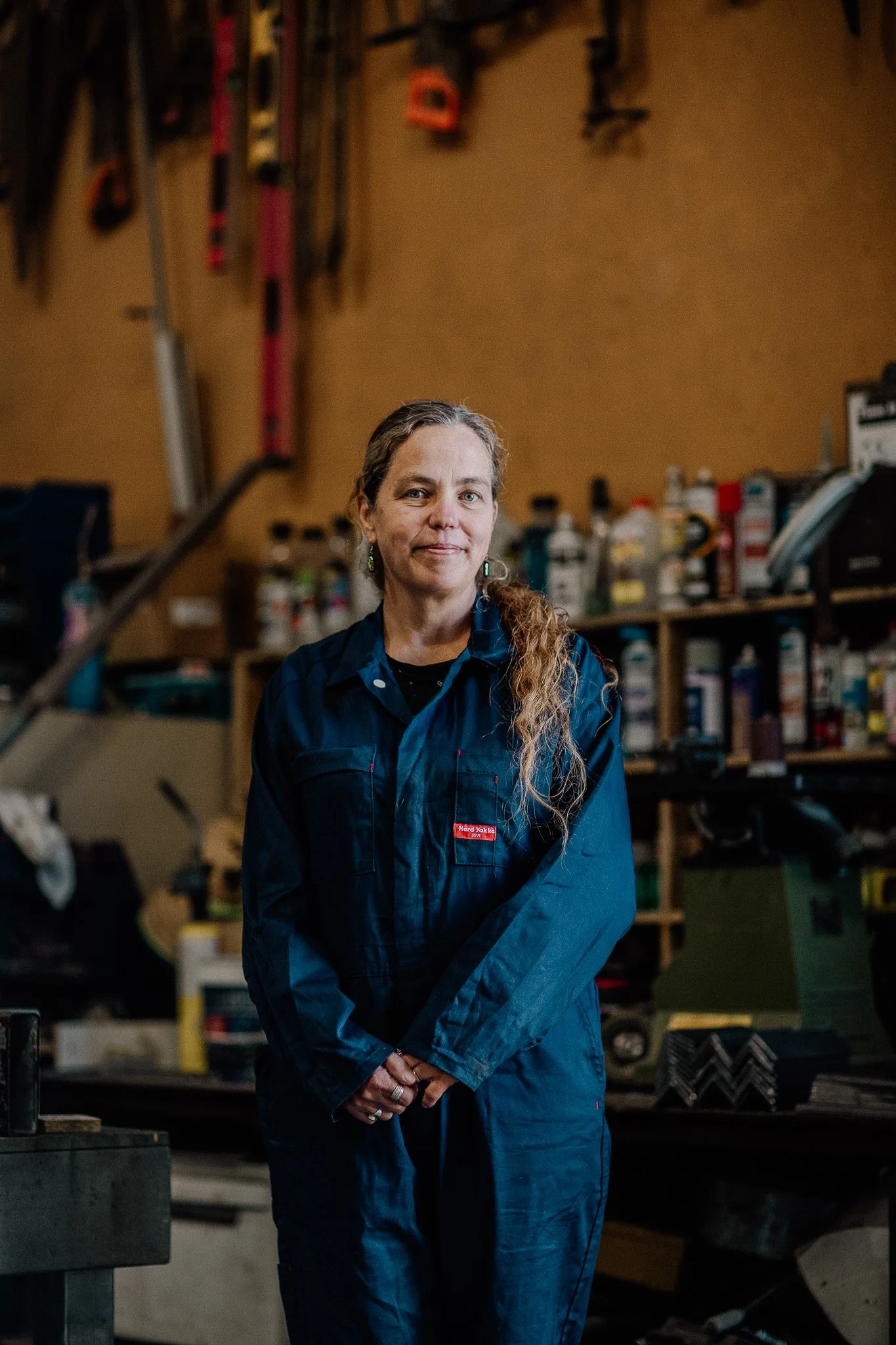 A woman with long, wavy hair wearing earrings and a blue work shirt standing in a workshop surrounded by tools and supplies.