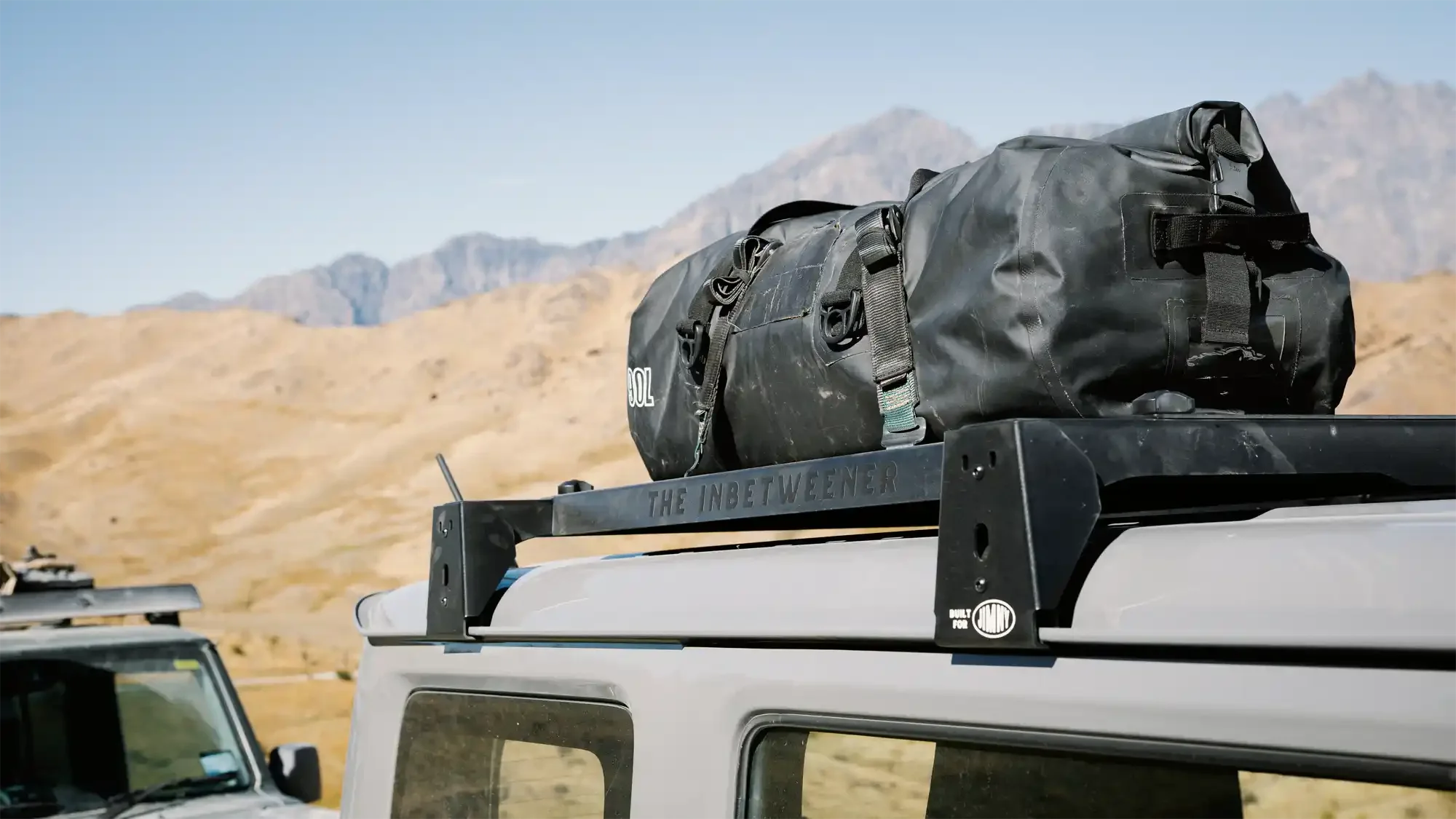 View of a black duffel bag secured on a rooftop cargo rack of a grey Jimny, set against a dry, mountainous landscape with a clear blue sky.