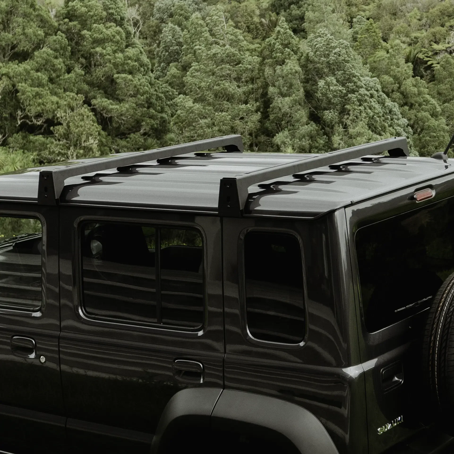 Side view of a charcoal Jimny 5-door with two black roof bars in front of NZ native bush.