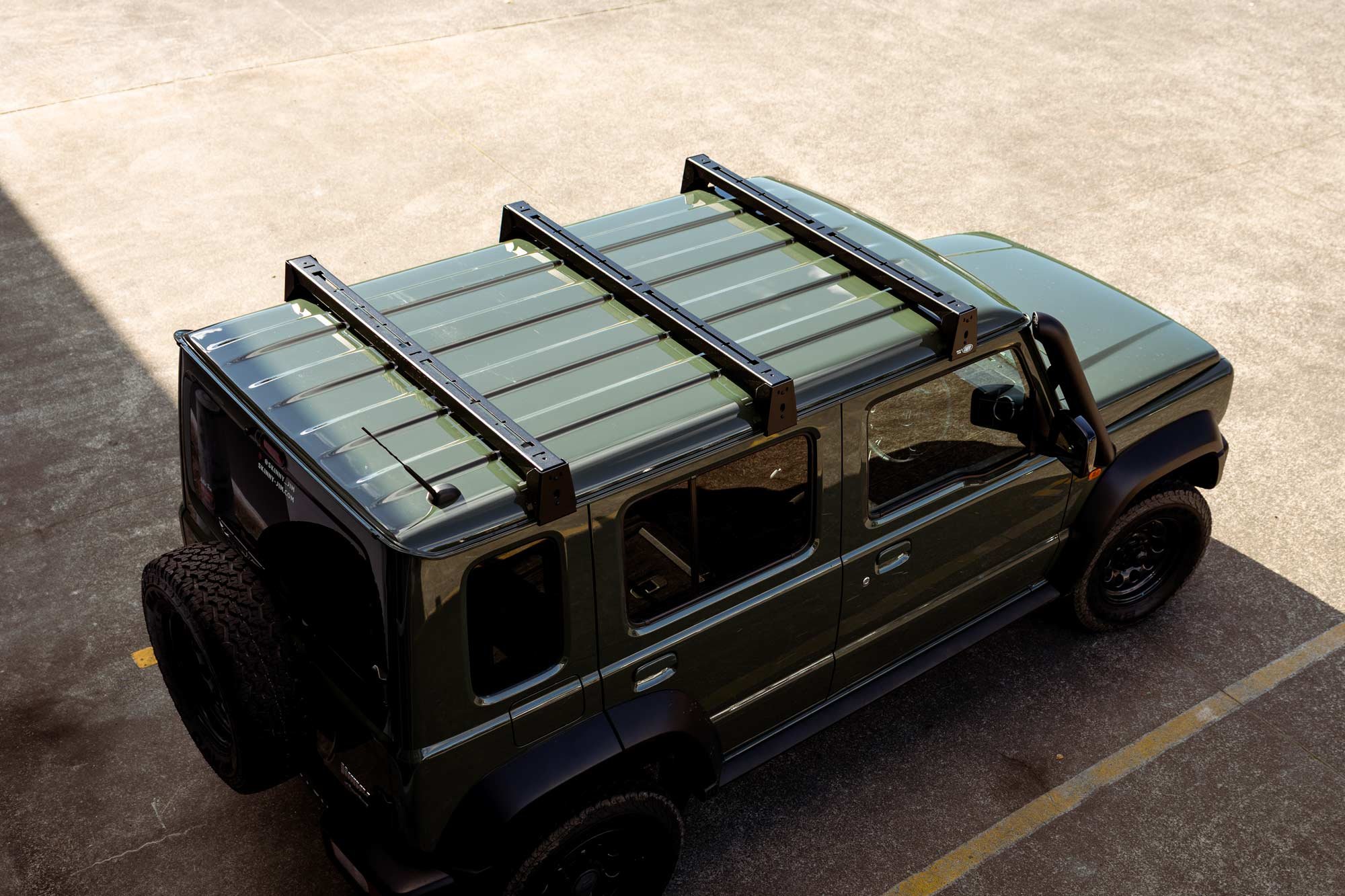 Jimny 5-Door with three black cross bars on roof, parked in outdoor parking lot, viewed from above