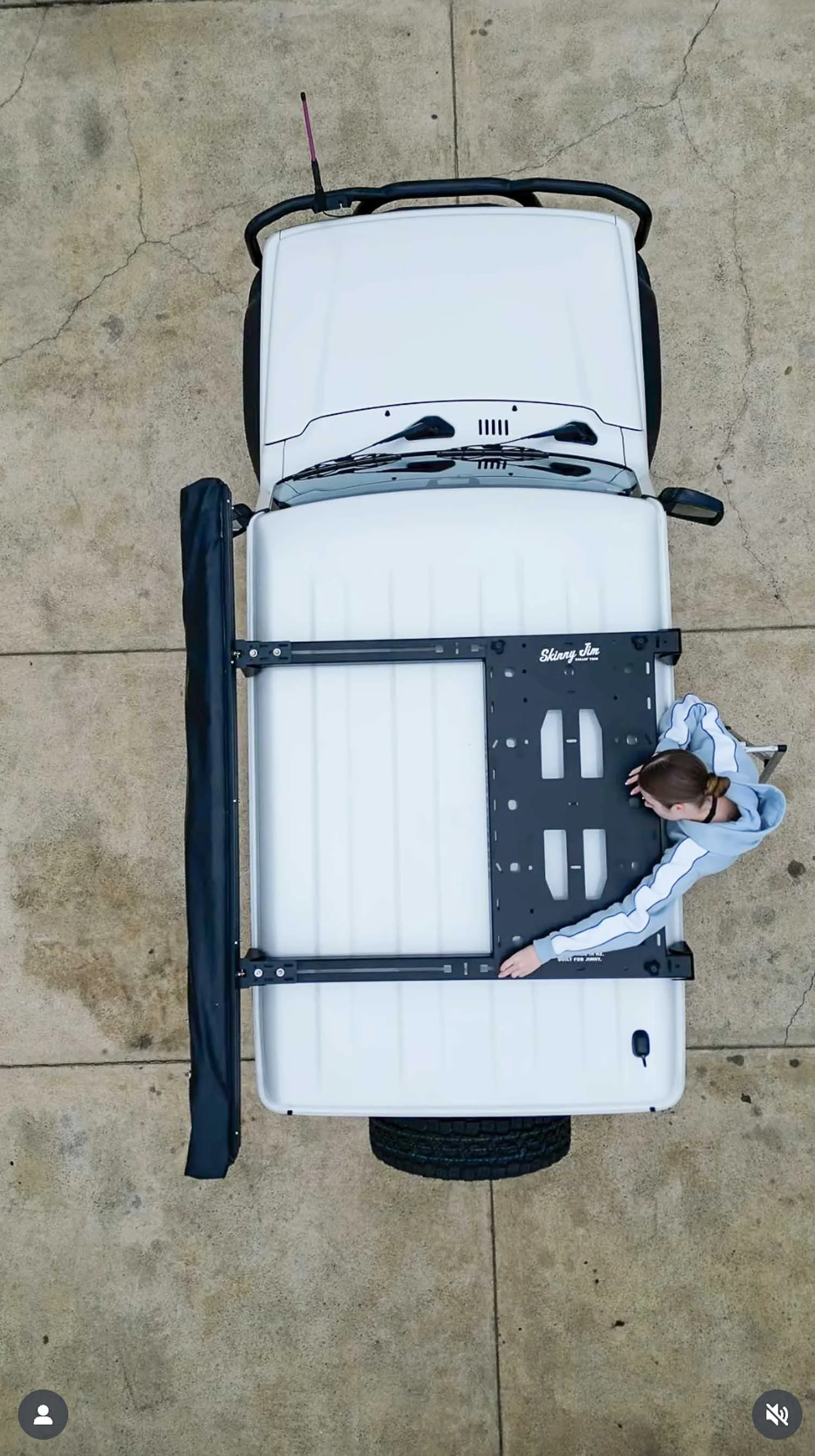 Top-down view of a white Suzuki Jimny with a roof rack and a woman installing a roof rack on the vehicle's roof.