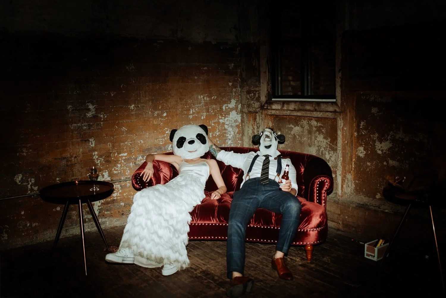 Bride and groom in playful animal masks lounging on a velvet couch at their reception.