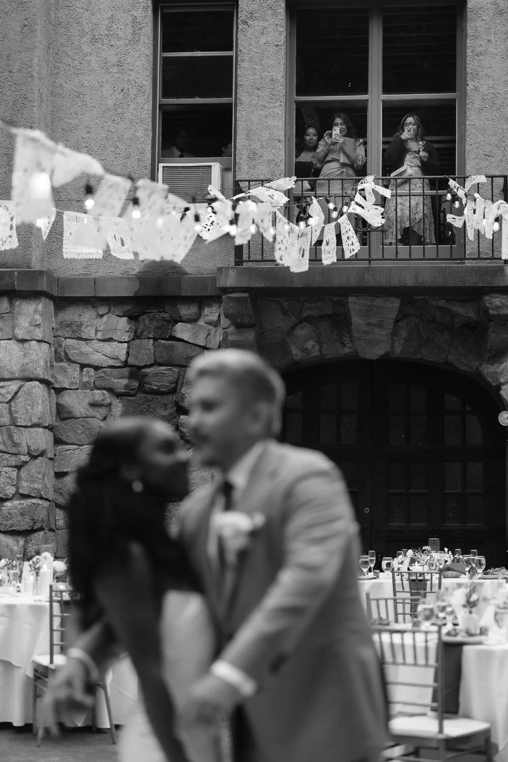 A couple dancing at a wedding reception with tables and chairs in the background, and people watching from a balcony above.