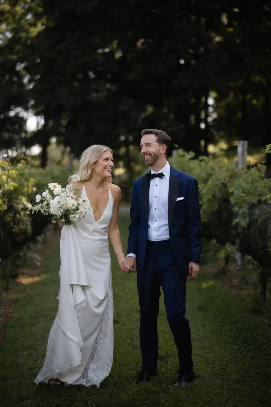 Just married couple stands in a vineyard sharing a laugh.