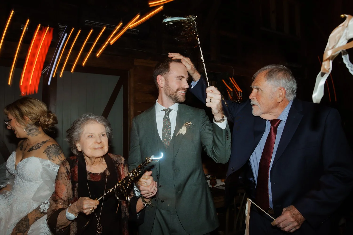 People celebrating at a wedding reception with sparklers, including a man in a gray suit and a woman with tattoos, in a dimly lit indoor venue.