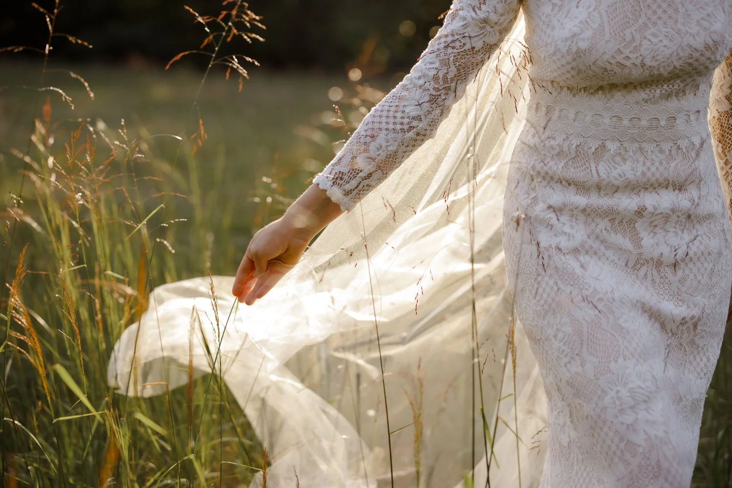 A person in a white lace dress standing in a grassy field, holding a sheer fabric in their hand as the sunlight shines through.