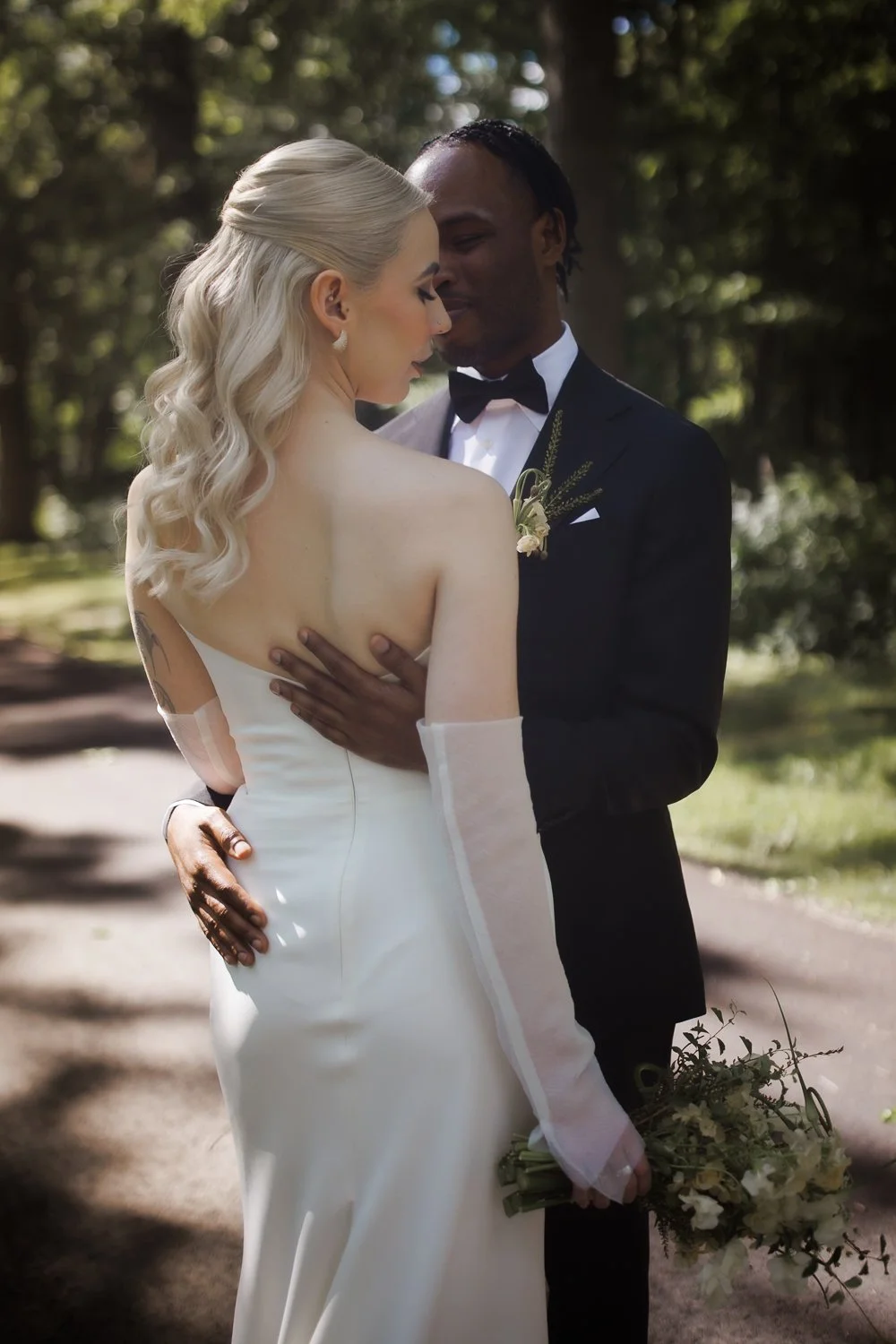 A wedding couple, with the bride in a white gown with sheer sleeves and the groom in a black tuxedo with a bow tie, embracing outdoors with trees in the background. The bride holds a bouquet of flowers.