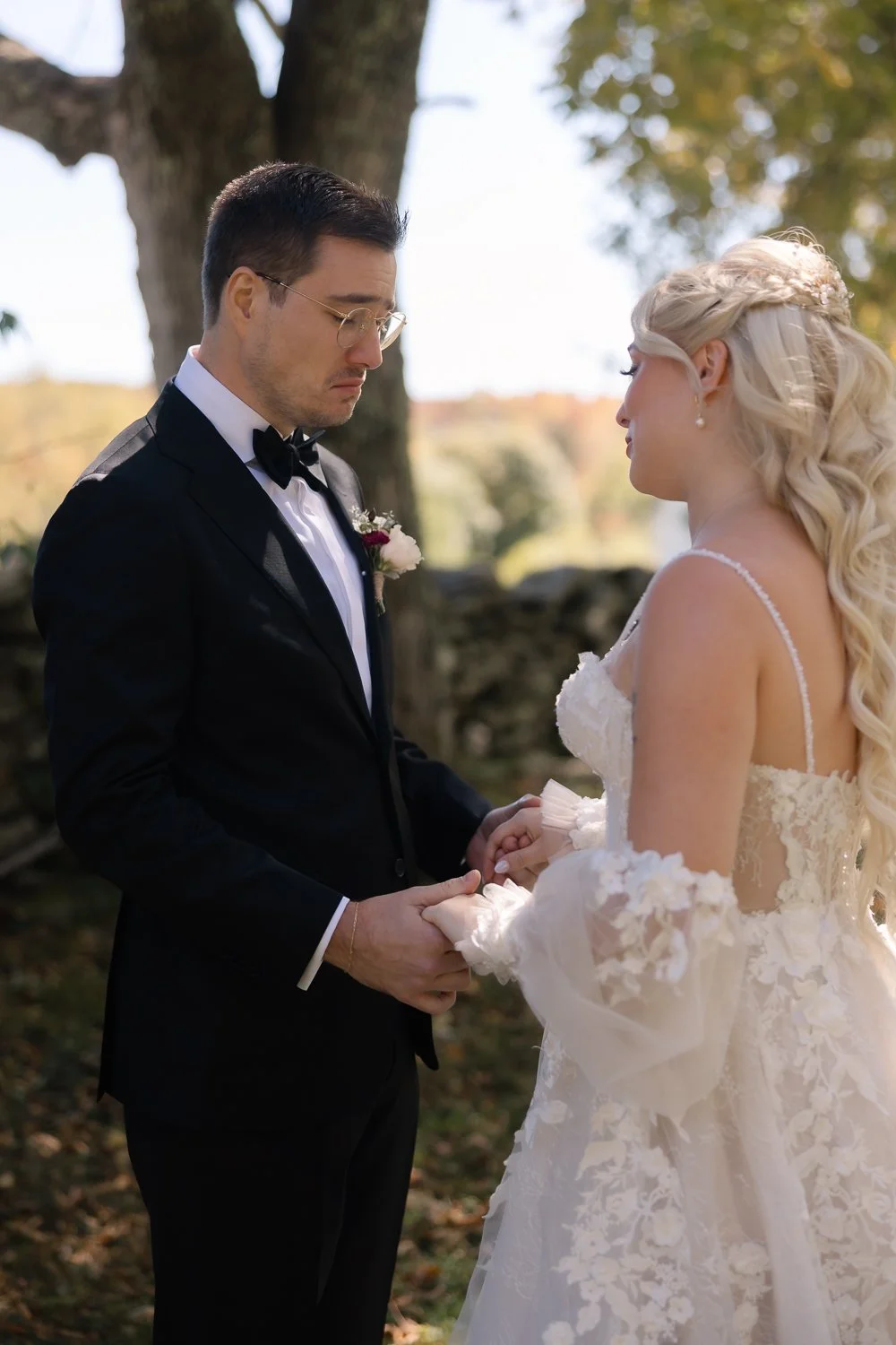 Groom tears up during a first look as the bride in a braided half-up style and lace gown holds his hands beneath a large tree.