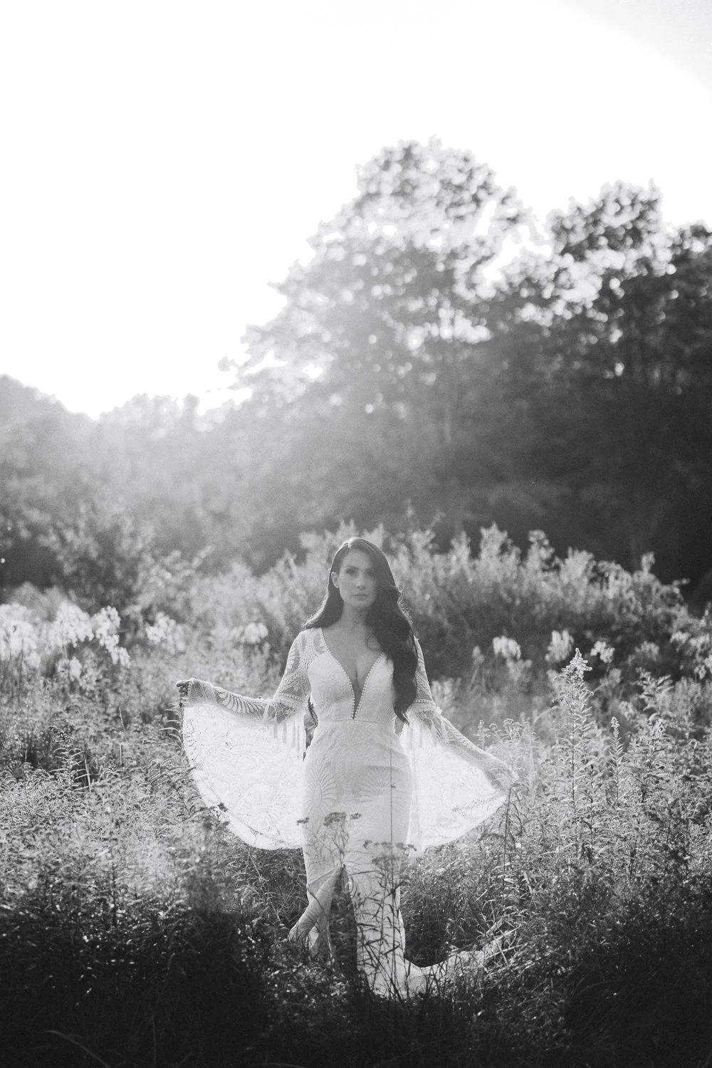 Editorial black and white portrait of a bride in a lace gown with bell sleeves standing in a sunlit field of wildflowers near Rhinebeck.