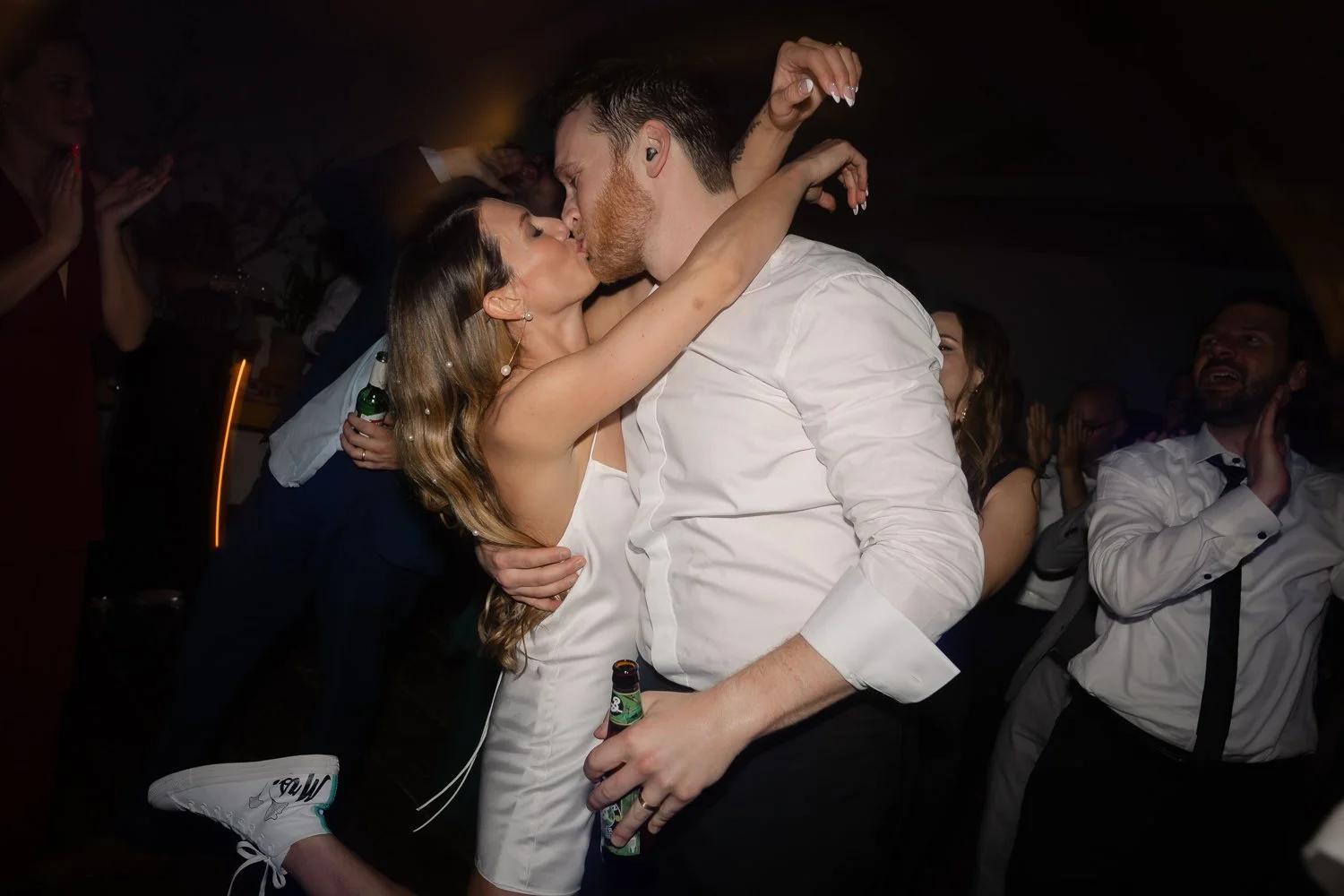 Bride in a short white dress and groom in a white shirt kissing on a crowded dance floor with beers in hand.