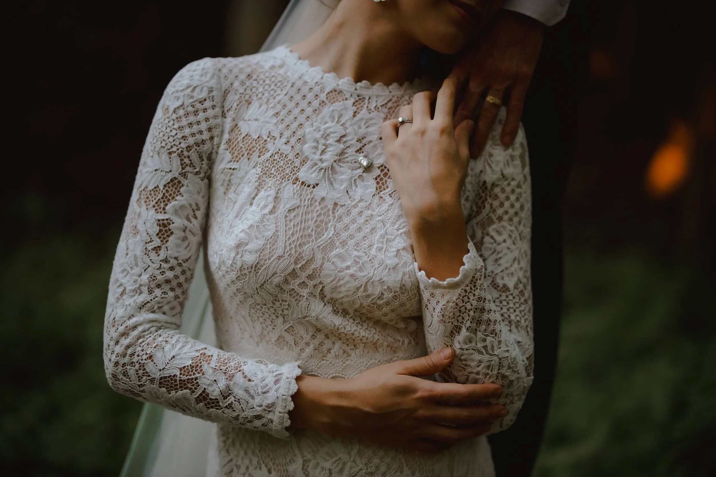 Detail of a bride's lace sleeve and engagement ring resting against her chest.