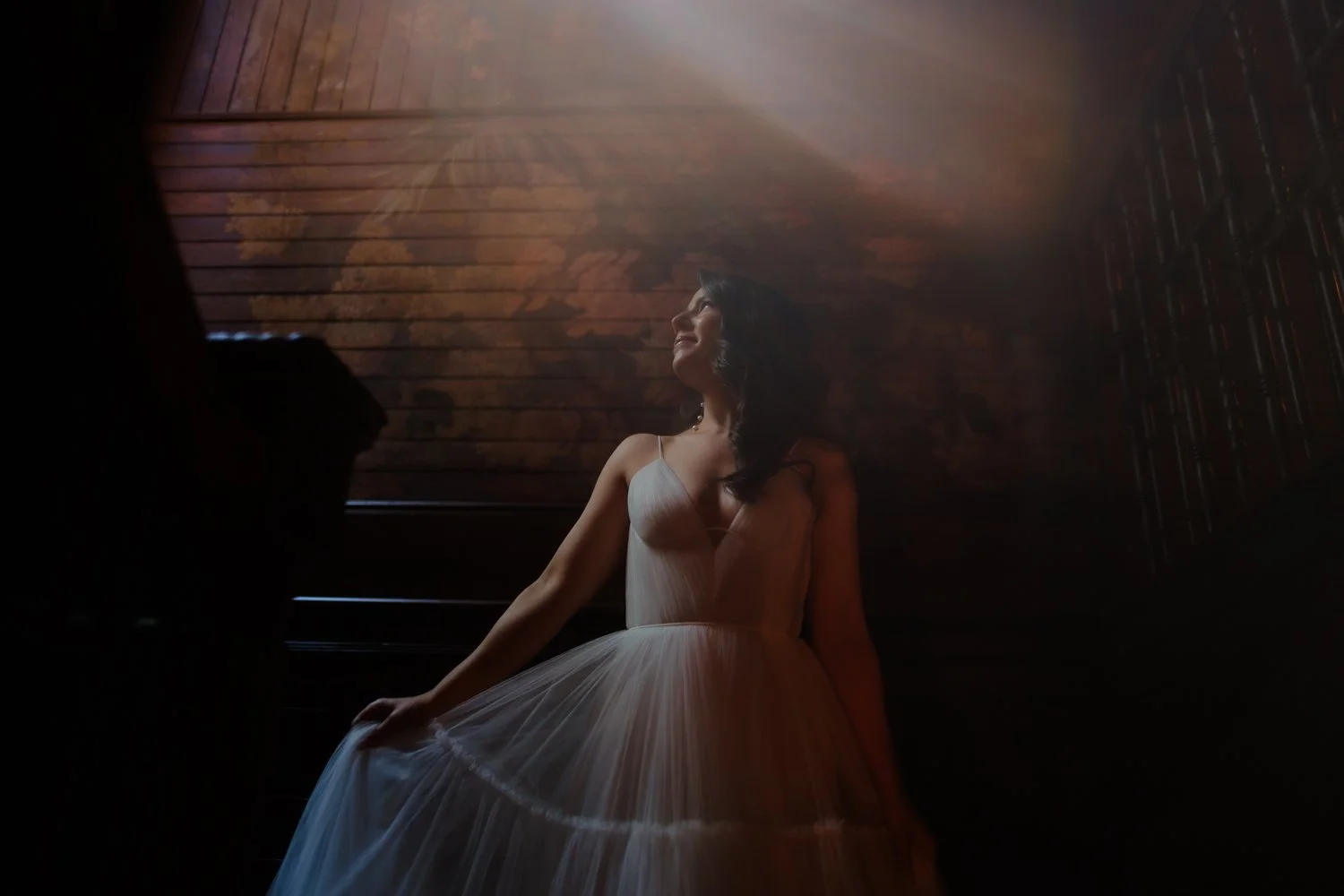Woman in a white wedding dress standing indoors with soft lighting and wooden wall background.