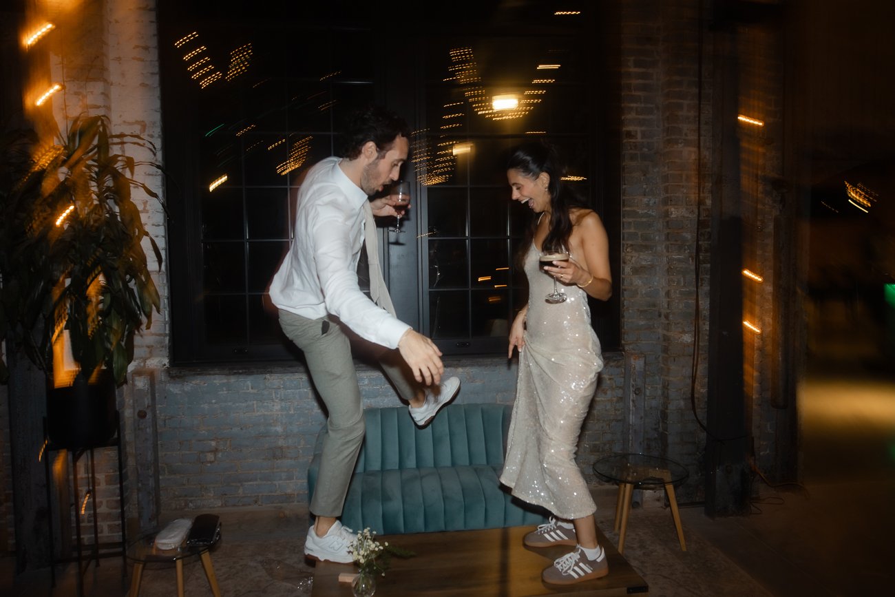 Couple dances on a table while holding drinks with lights swirling around them during a wedding.