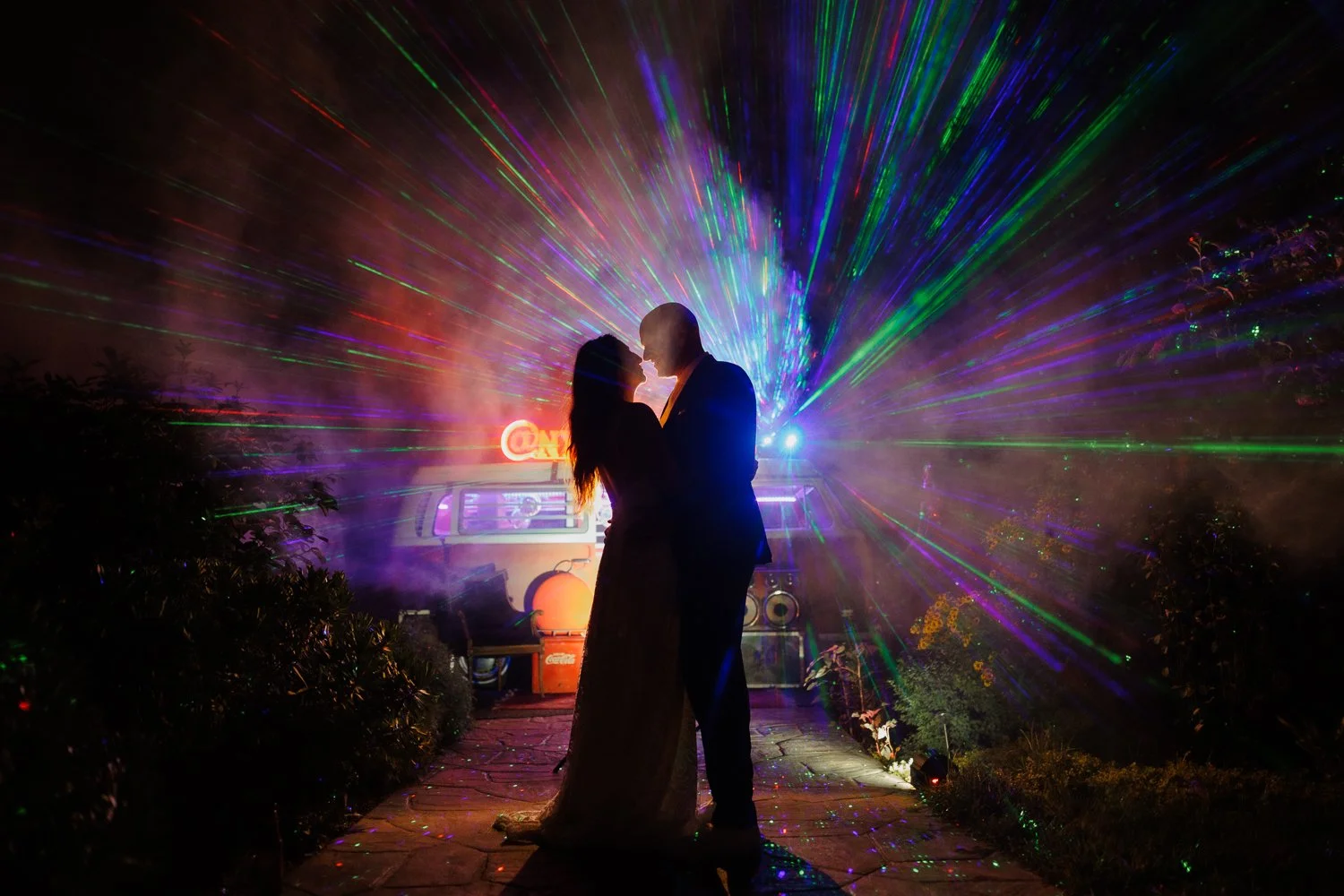 Couple in silhouette about to kiss on a stone path as colorful laser beams and smoke glow behind a vintage camper van.