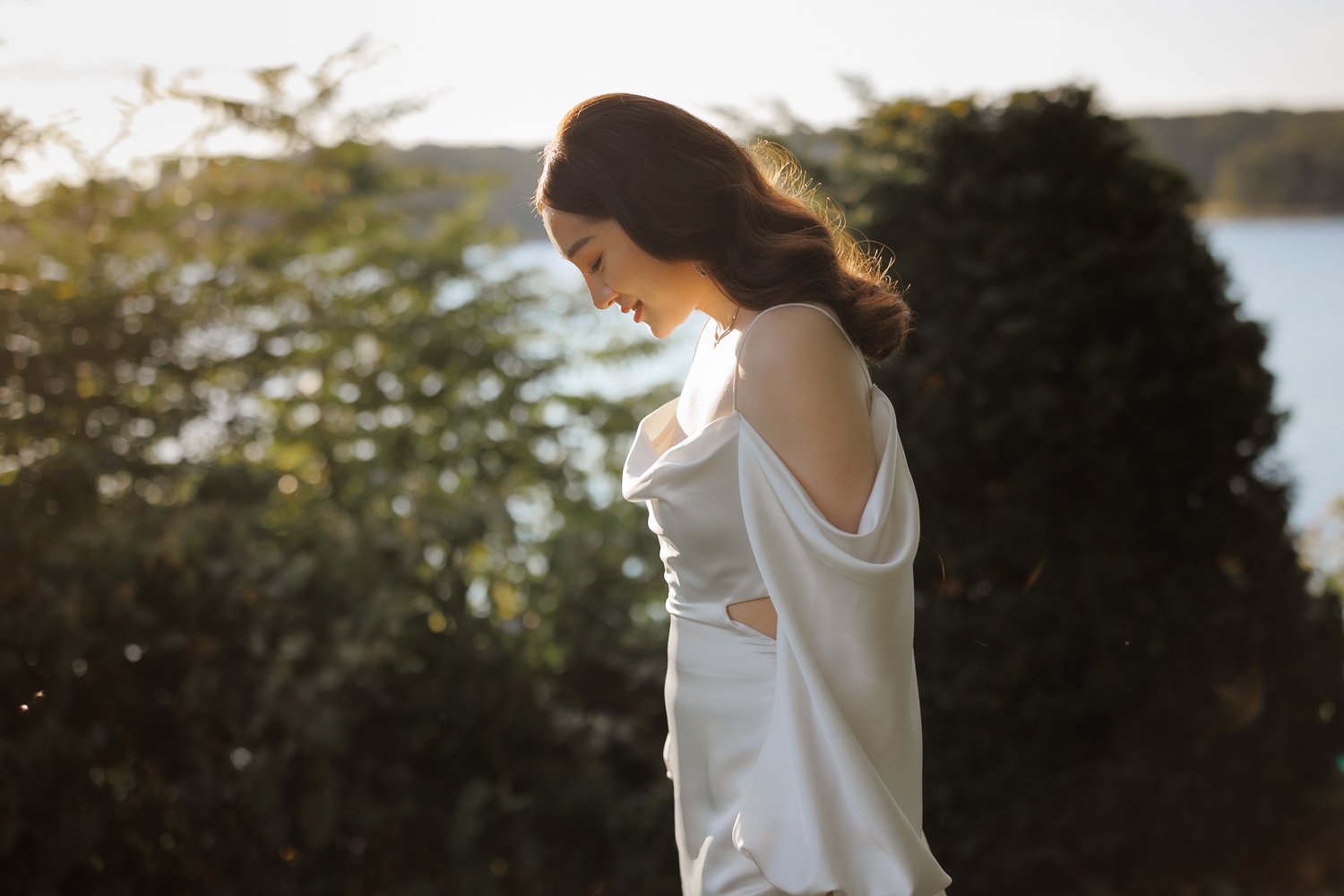 Bride in a satin gown with a draped sleeve smiling down in warm backlight with a lake behind her.