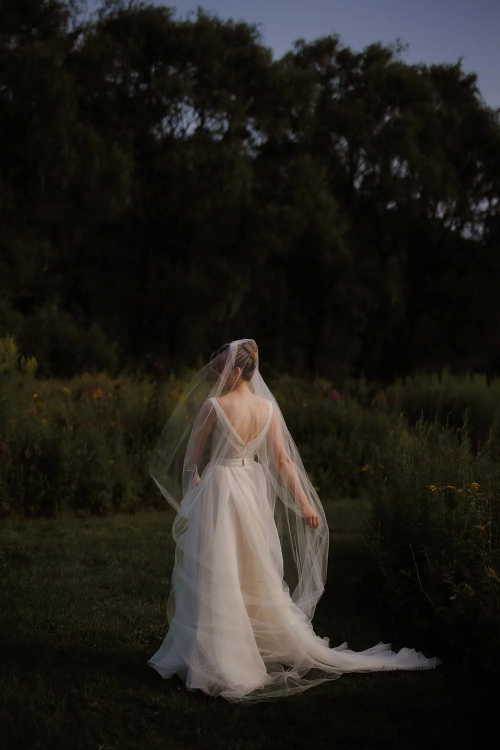 Bride in a long tulle veil and gown facing a field of wildflowers at twilight.