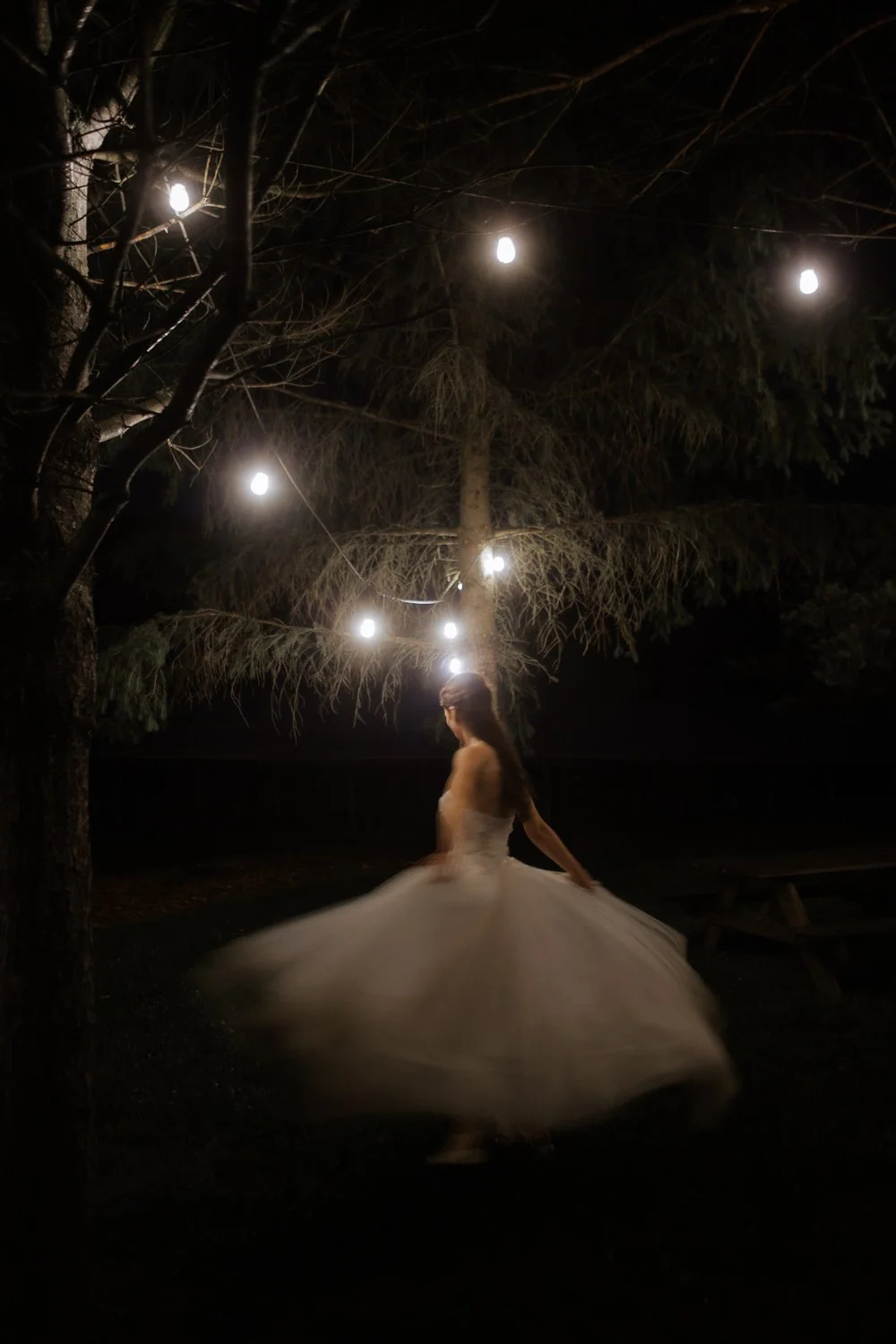 Bride twirling her gown under string lights strung through the trees at night.