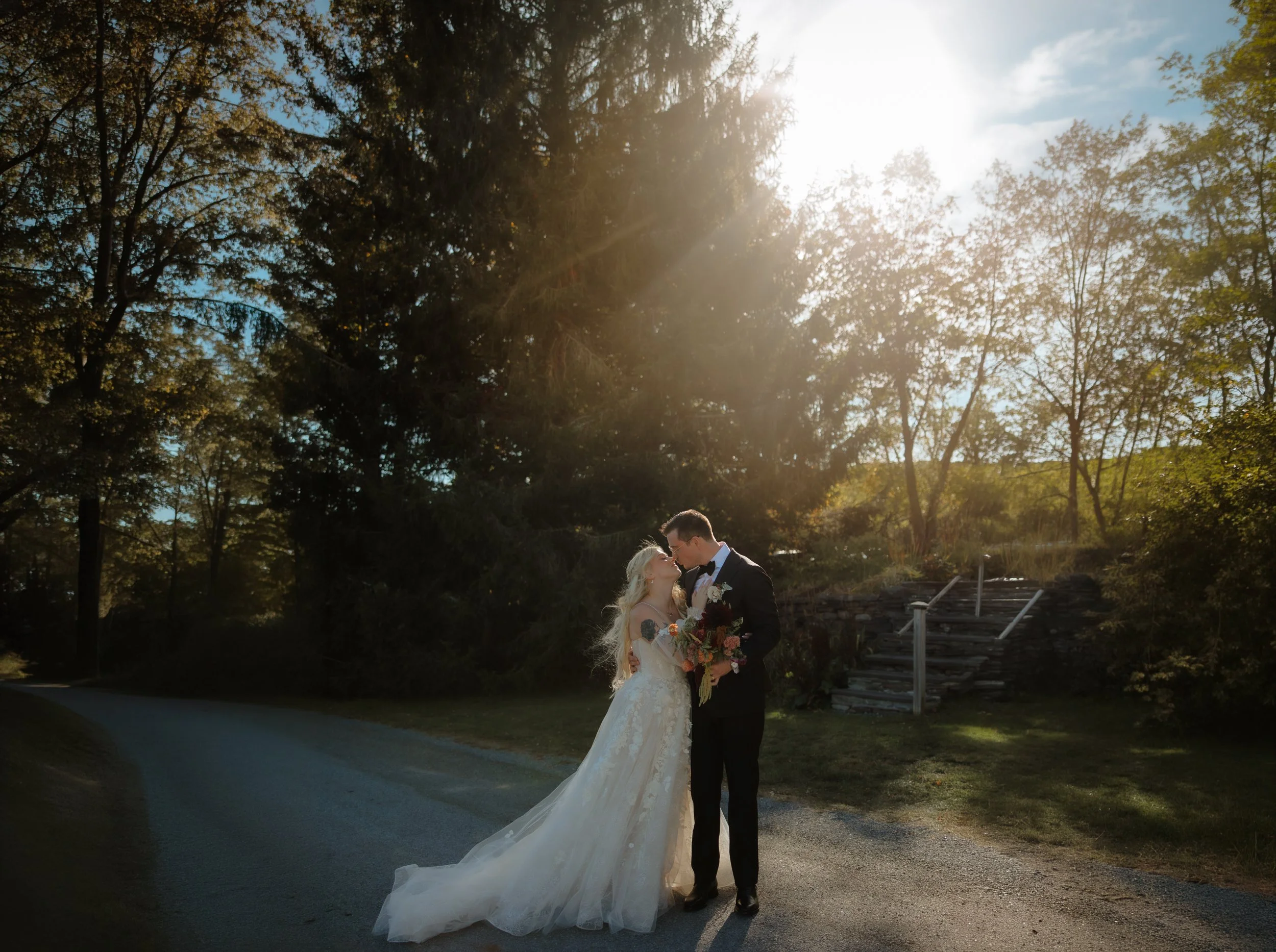Bride and groom kissing on a sunlit path lined with trees at golden hour.