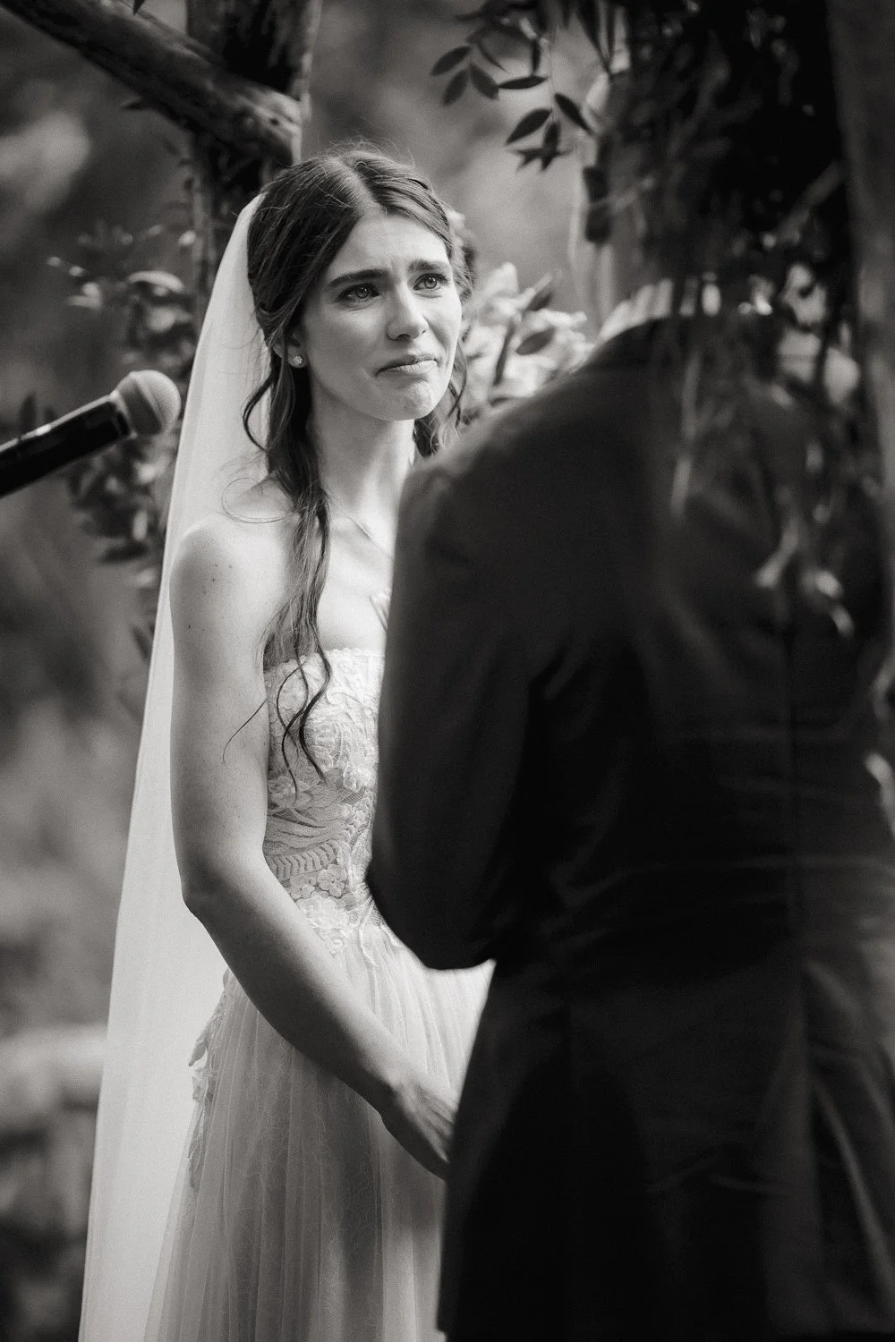 A bride tears up during her wedding vows, standing in front of her groom, with a blurred background of trees and plants.