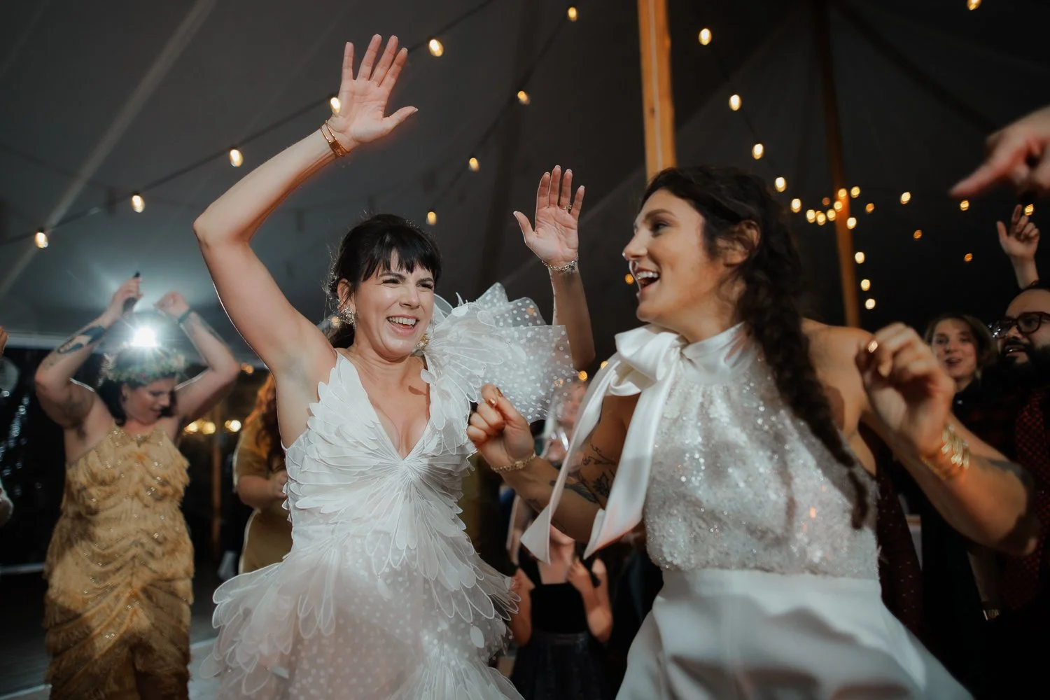 Two women dancing and smiling at a party under string lights, one in a white dress with ruffled sleeves, the other in a white halter top with a bow.