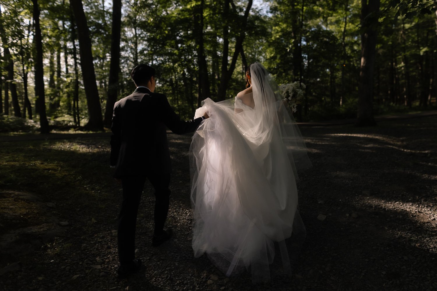 Groom carries brides veil through a pocket of light for a moody candid.