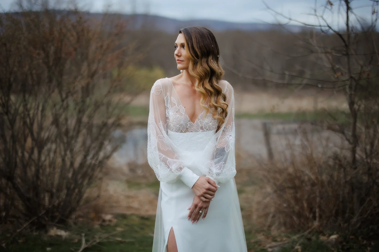 A woman in a white dress with lace details, standing outdoors by a pond and bare bushes, during overcast weather.