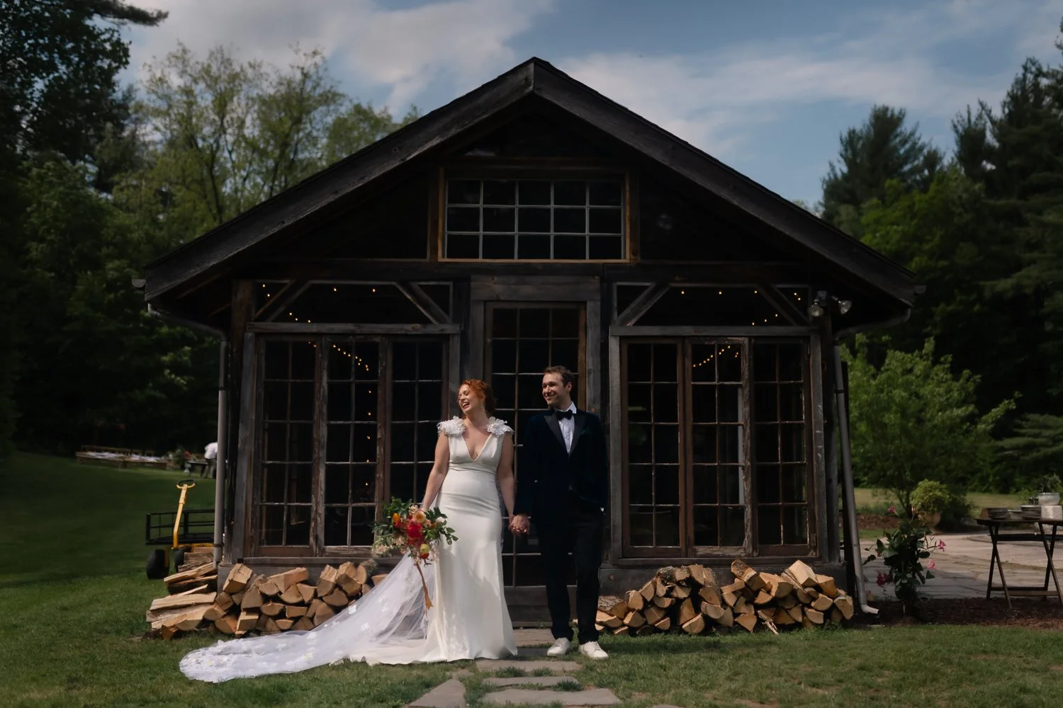Bride in a satin gown and groom in a tuxedo holding hands in front of Foxfire's greenhouse with stacked firewood, her long veil trailing across the grass.