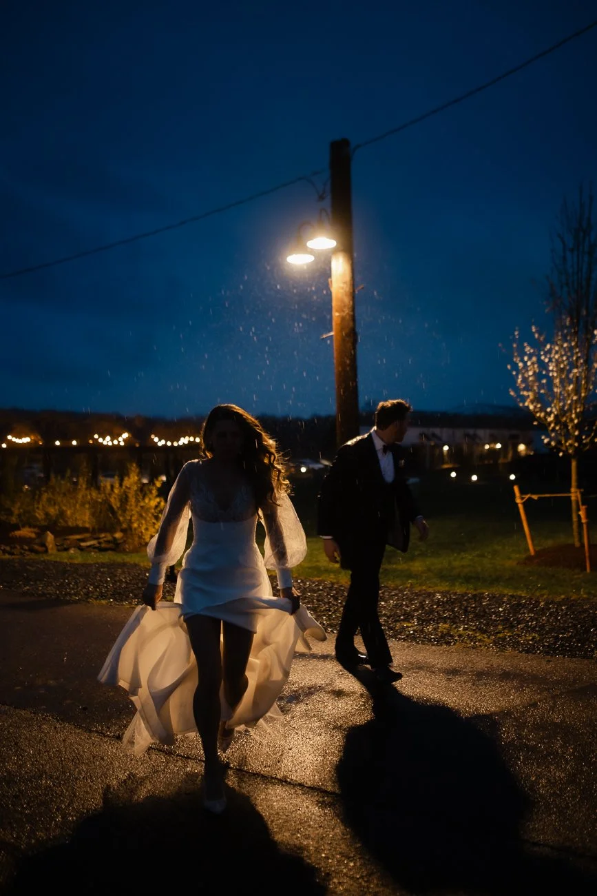 Bride and groom are silhouetted against the blue night sky under a yellow street light that shows the rain coming down.