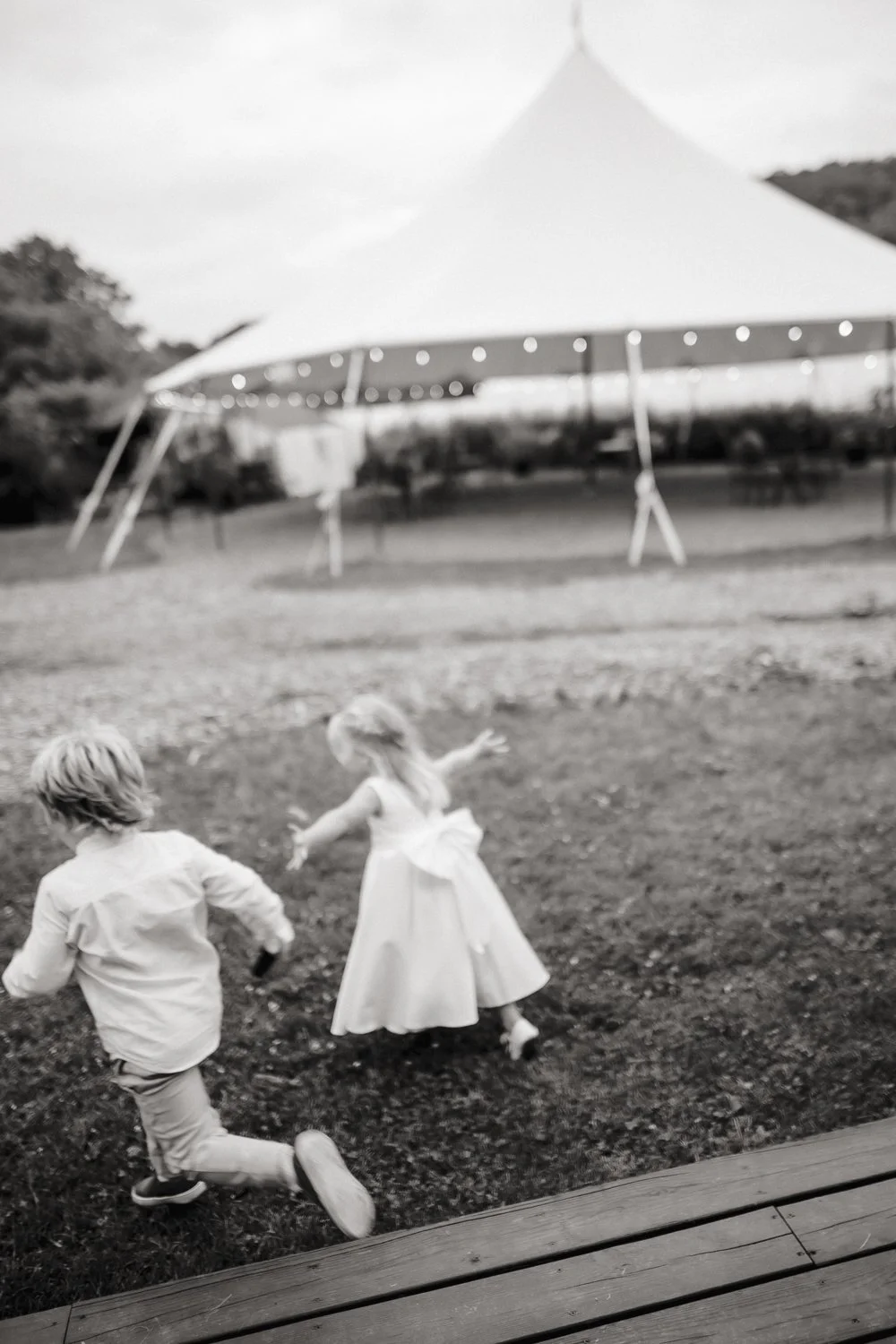 Two children, a boy and a girl, running on grass near a wooden deck at an outdoor event with a large white tent in the background.