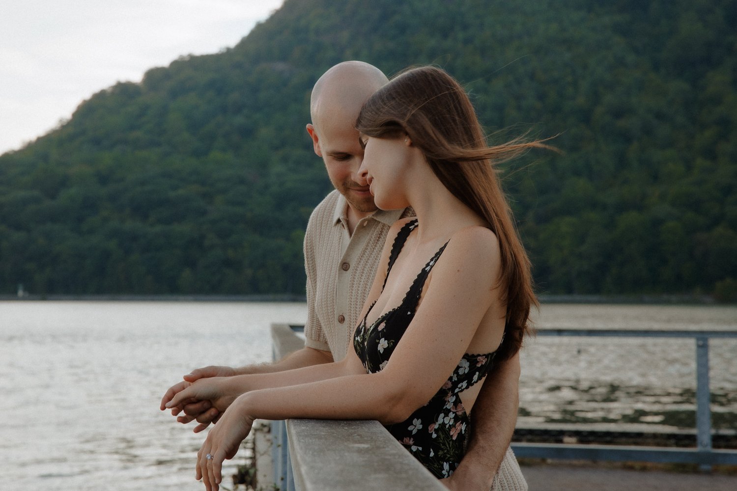 Engaged couple leaning against a railing overlooking the Hudson River with mountains in the background.