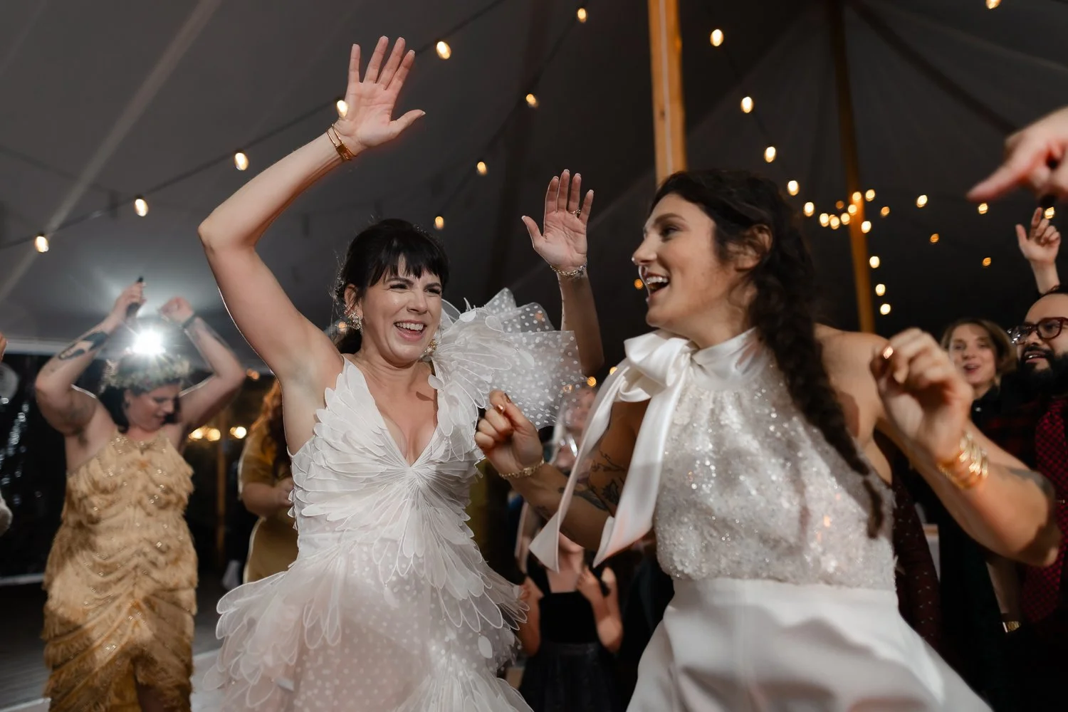 Two women dancing and smiling at a lively party under string lights, with others dancing around them.