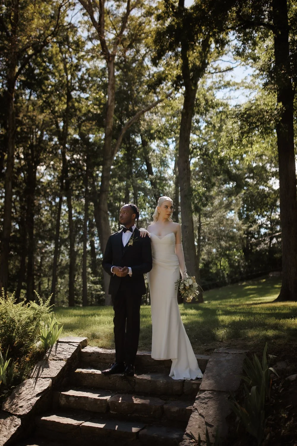 A bride and groom walk down stone steps in a forested area, with tall trees and sunlight filtering through the leaves in the background.
